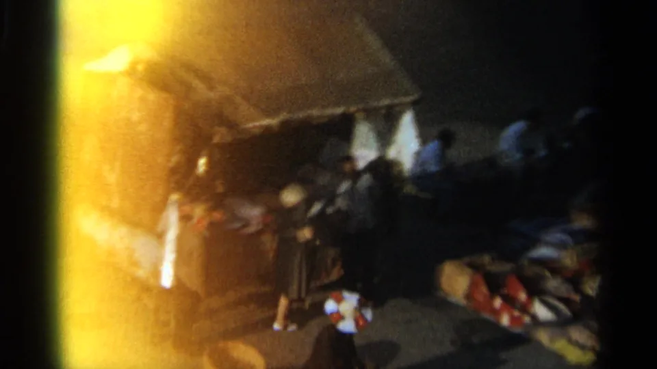 Closer grainy frame from 1965 Tangier showing a small market kiosk, nearby people and merchandise at dusk.