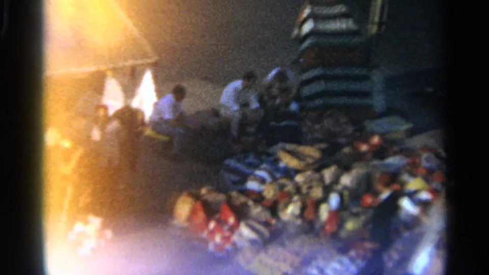 1965 Tangier night market frame showing several people near piles of textiles and bags, with bright flare at the edge of the 8mm frame.