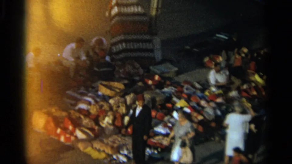 Tangier, Morocco night market in 1965, with several adults around colorful textiles and bags in a warmly lit outdoor stall area.