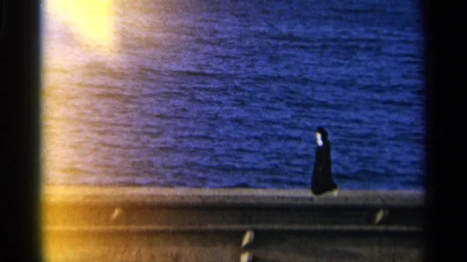 Tangier, Morocco 1965 frame showing a woman in a dark religious habit walking along a seaside promenade with blue water beyond.