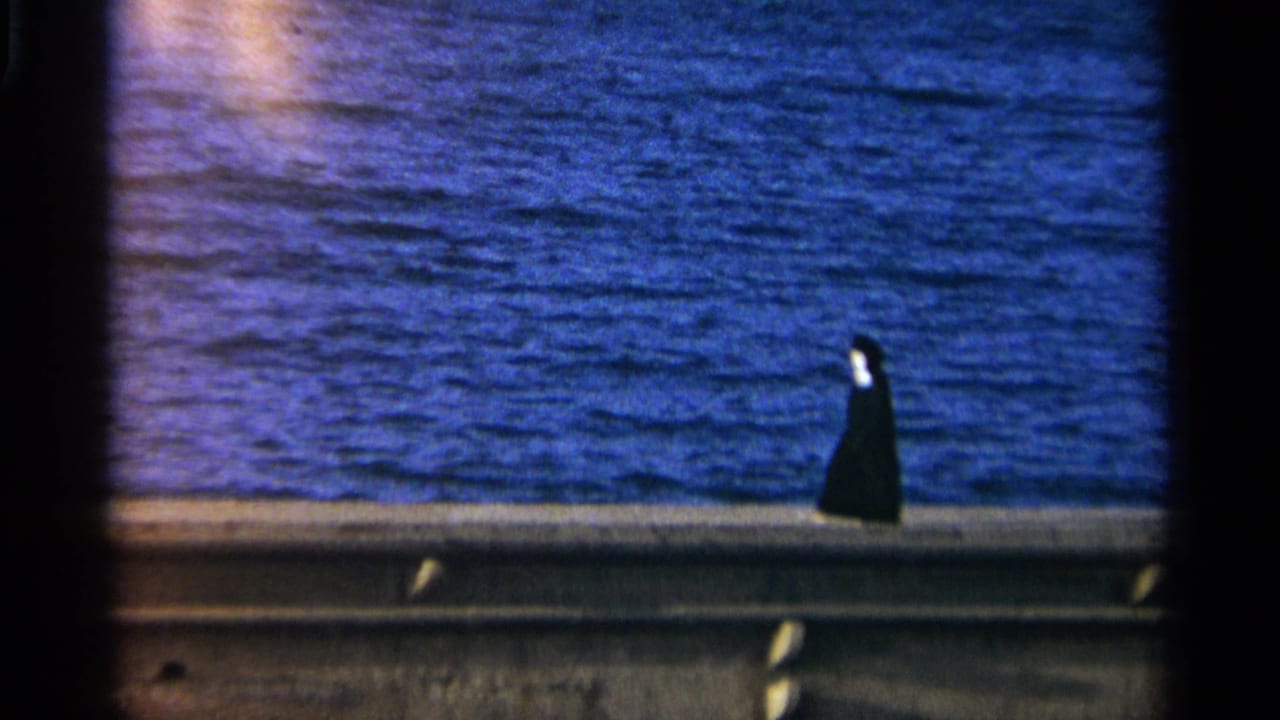 A distant figure in black walks along a concrete waterfront ledge in Tangier, Morocco, in 1965 home-movie footage.