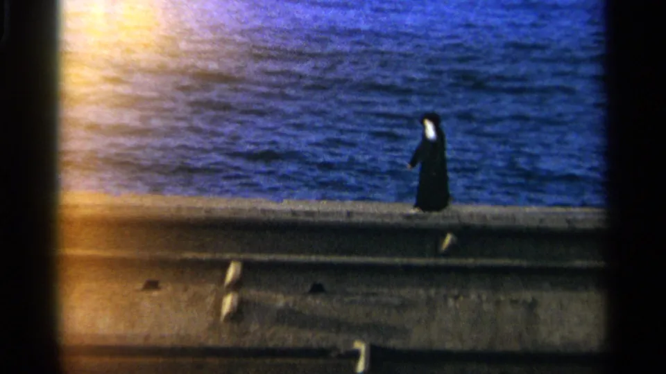 A distant pedestrian in black continues leftward along the Tangier waterfront, framed by grainy 8mm color and blue water.