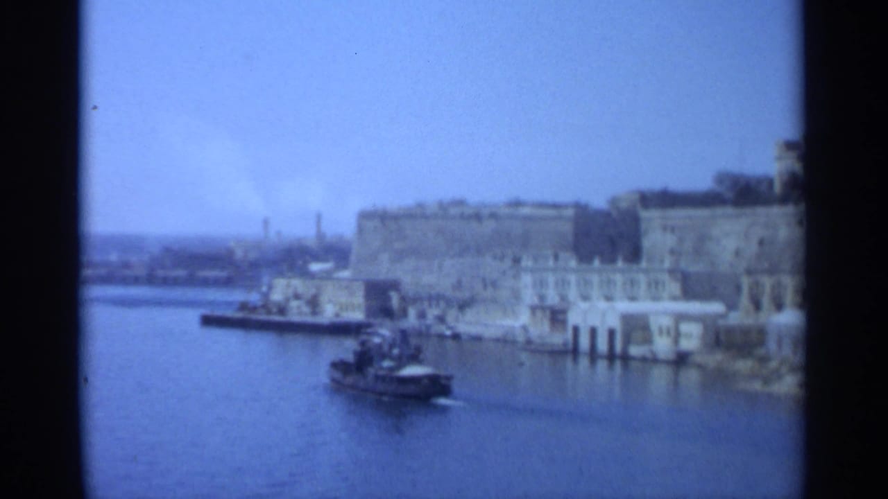 1965 Tangier waterfront view with stone harbor fortifications, pale buildings, calm water, and a small boat near shore.