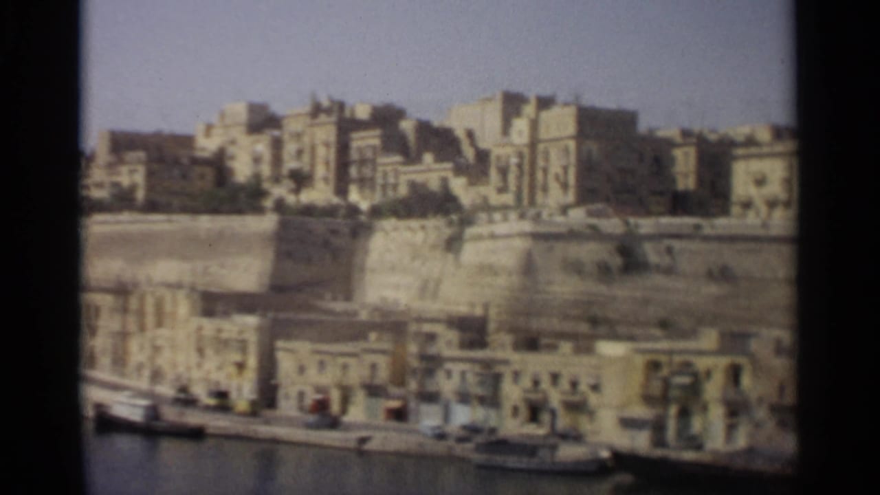 1965 home-movie frame of Tangier waterfront architecture, with harbor water, quay buildings, and dense structures above.