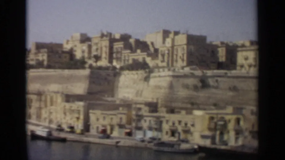 1965 home-movie frame of Tangier waterfront architecture, with harbor water, quay buildings, and dense structures above.
