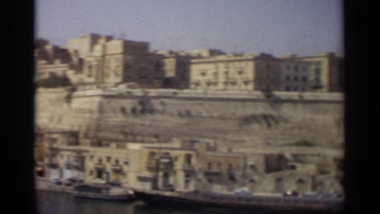 Wide 1965 Tangier waterfront view with calm harbor, stone quay, moored boats, and sunlit buildings in soft 8mm focus.
