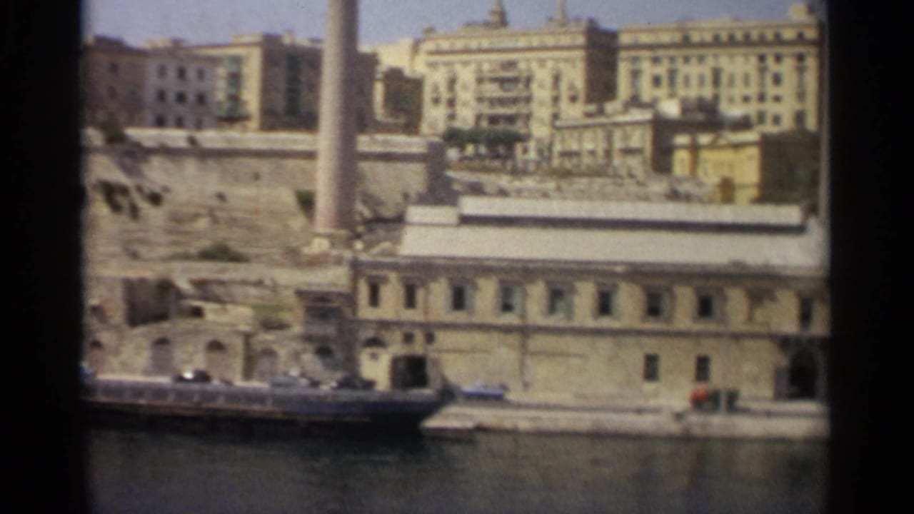 Tangier harbor frontage in 1965 with a long low building, boats along the water, hillside masonry, and pale urban blocks behind.