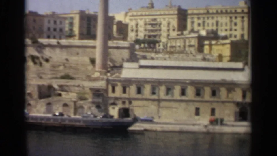 Tangier harbor frontage in 1965 with a long low building, boats along the water, hillside masonry, and pale urban blocks behind.