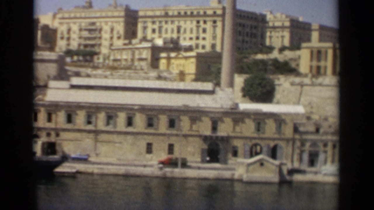 Soft 8mm frame of Tangier's waterfront with arched doorways, a small boat, water in the foreground, and pale buildings above.