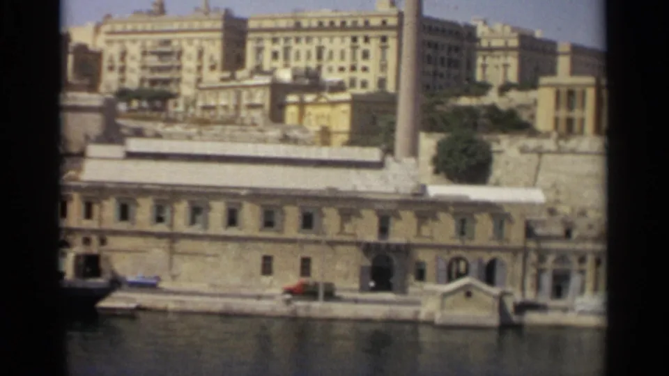 Soft 8mm frame of Tangier's waterfront with arched doorways, a small boat, water in the foreground, and pale buildings above.