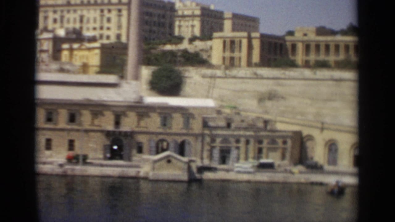 1965 Tangier harbor view showing a long waterfront structure, stone wall, small boats, water, and pale city buildings beyond.