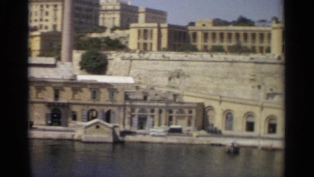 Waterfront buildings and a stone wall above a quay with moored boats and parked cars.