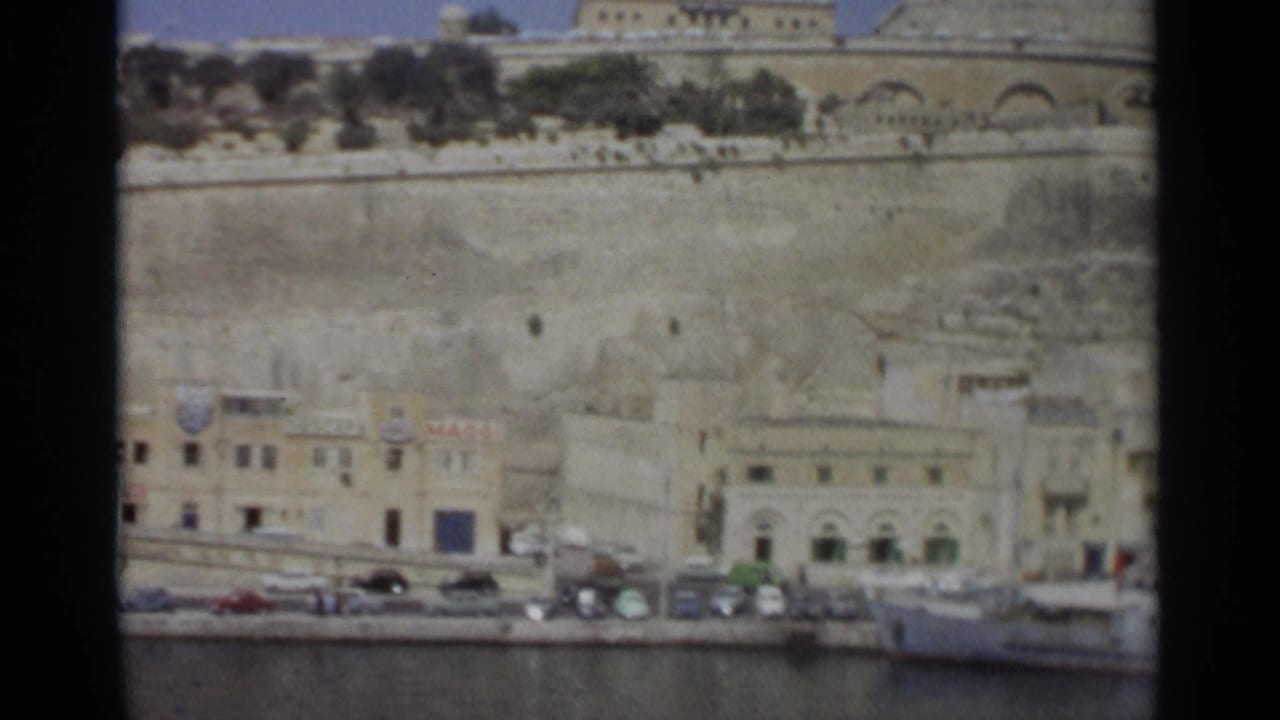 Wide 1965 Tangier harborfront view showing a waterfront street, parked cars, stone retaining wall, arched building, and moored boat.