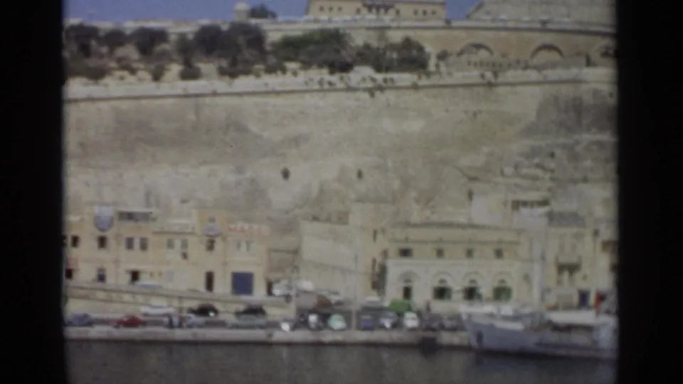 Wide 1965 Tangier harborfront view showing a waterfront street, parked cars, stone retaining wall, arched building, and moored boat.