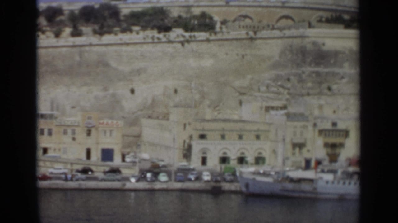 Waterfront buildings, parked cars, harbor water, and a docked white vessel below stone walls.