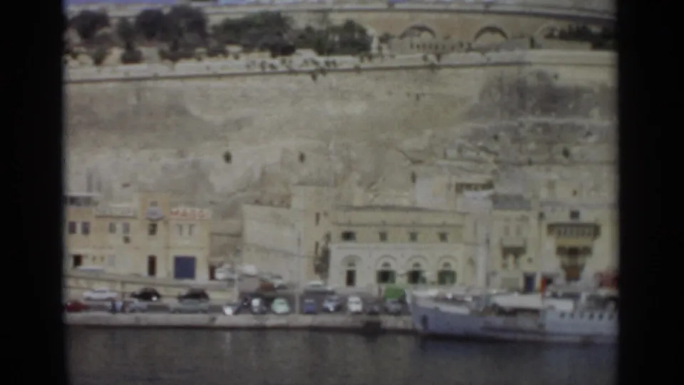 1965 Tangier waterfront view with a docked vessel, cars along the quay, pale buildings, and large stone fortification walls above the harbor.