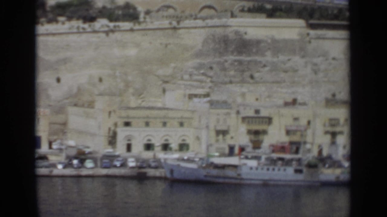 1965 Tangier harbor frame showing a white vessel at the quay, parked cars, arched waterfront facades, and weathered stone walls above town.