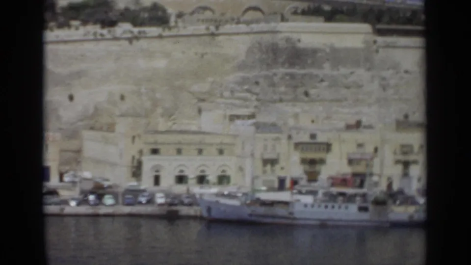 1965 Tangier harbor frame showing a white vessel at the quay, parked cars, arched waterfront facades, and weathered stone walls above town.