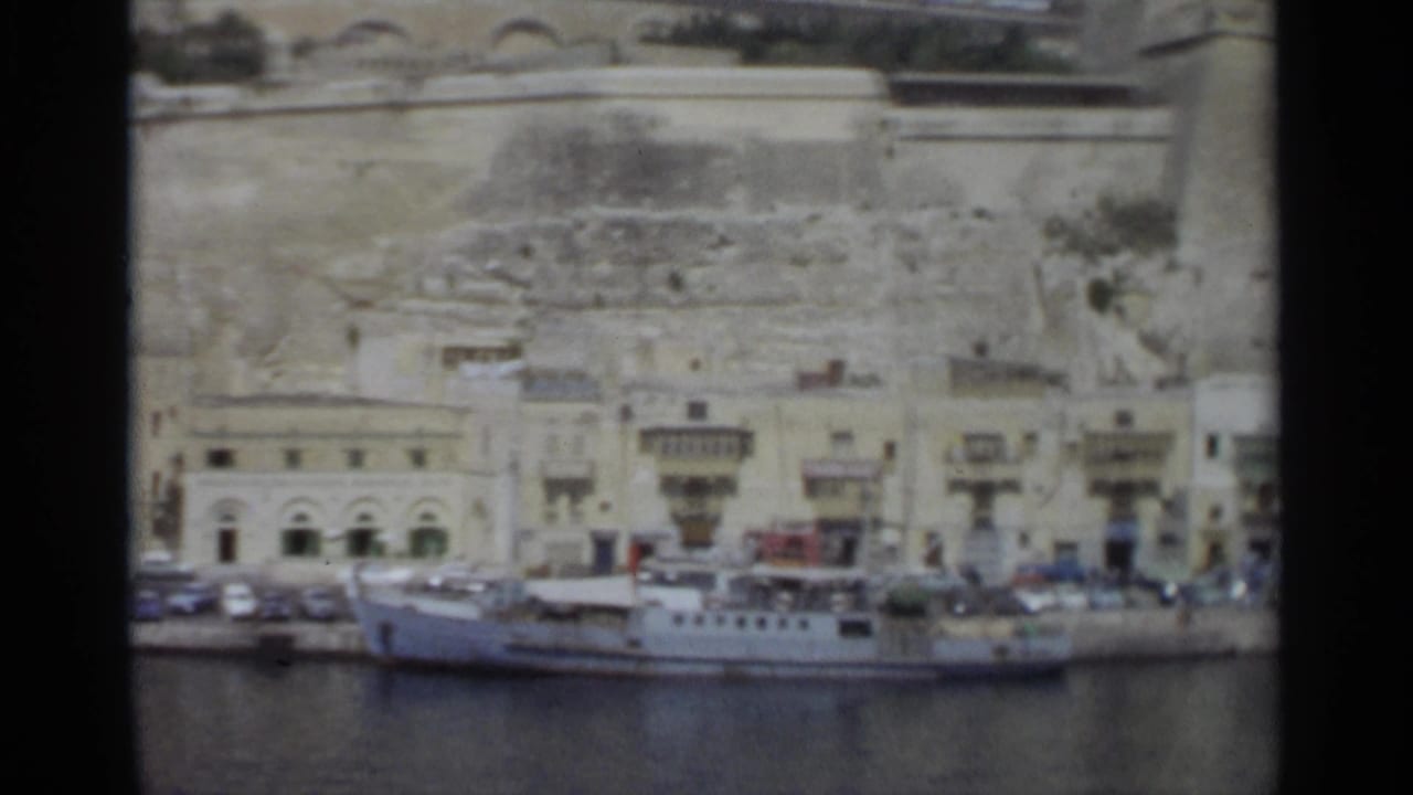 1965 Tangier waterfront seen from across the harbor, with the vessel lower frame, roadside cars, pale buildings, balconies, and stonework rising behind.