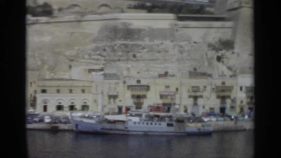 1965 Tangier waterfront seen from across the harbor, with the vessel lower frame, roadside cars, pale buildings, balconies, and stonework rising behind.