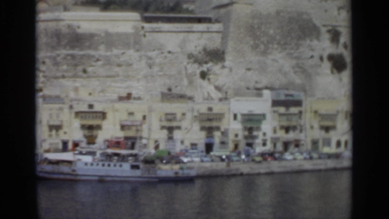 1965 Tangier shoreline frame with harbor water in front, a white vessel, a line of cars and buildings, and broad stone walls above the city edge.
