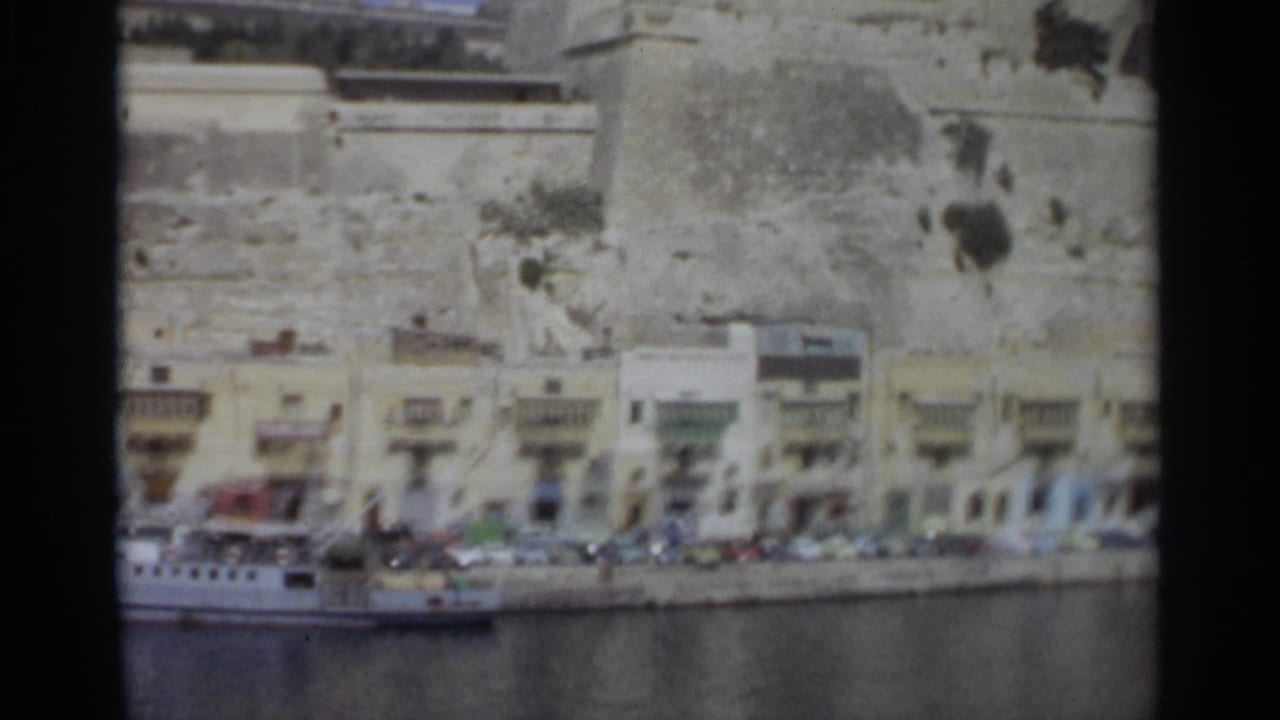 Waterfront street with parked cars, pale buildings, and a large stone wall above the road.