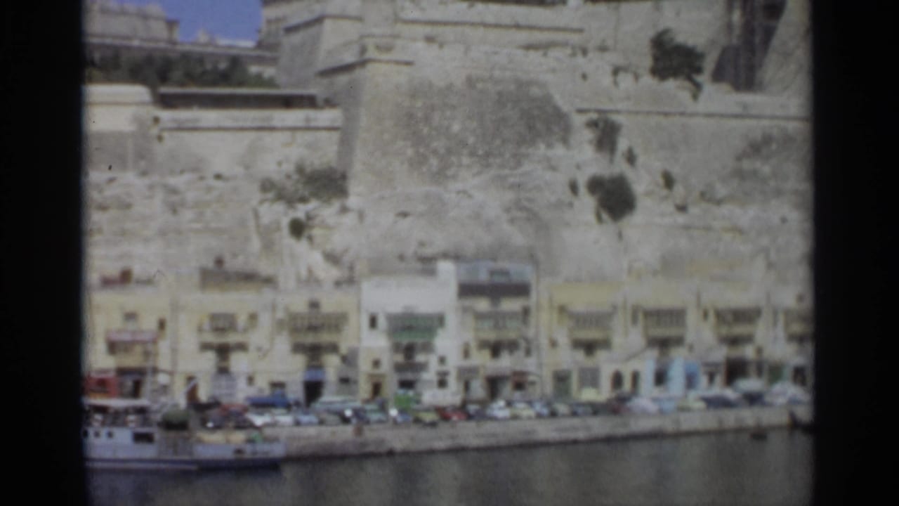 Tangier coastal street scene from the supplied 1965 footage, showing parked cars, waterfront buildings, and a fortress-like stone wall.
