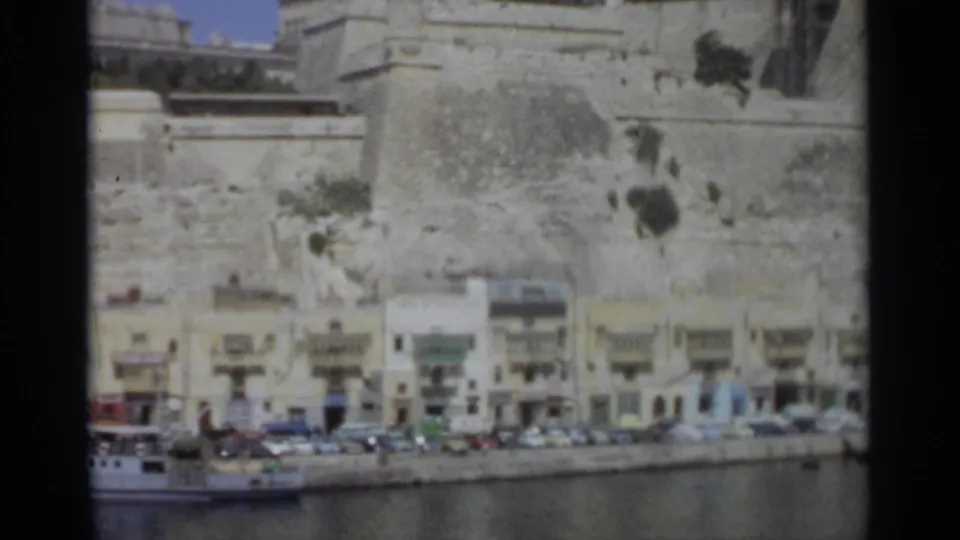 Tangier coastal street scene from the supplied 1965 footage, showing parked cars, waterfront buildings, and a fortress-like stone wall.