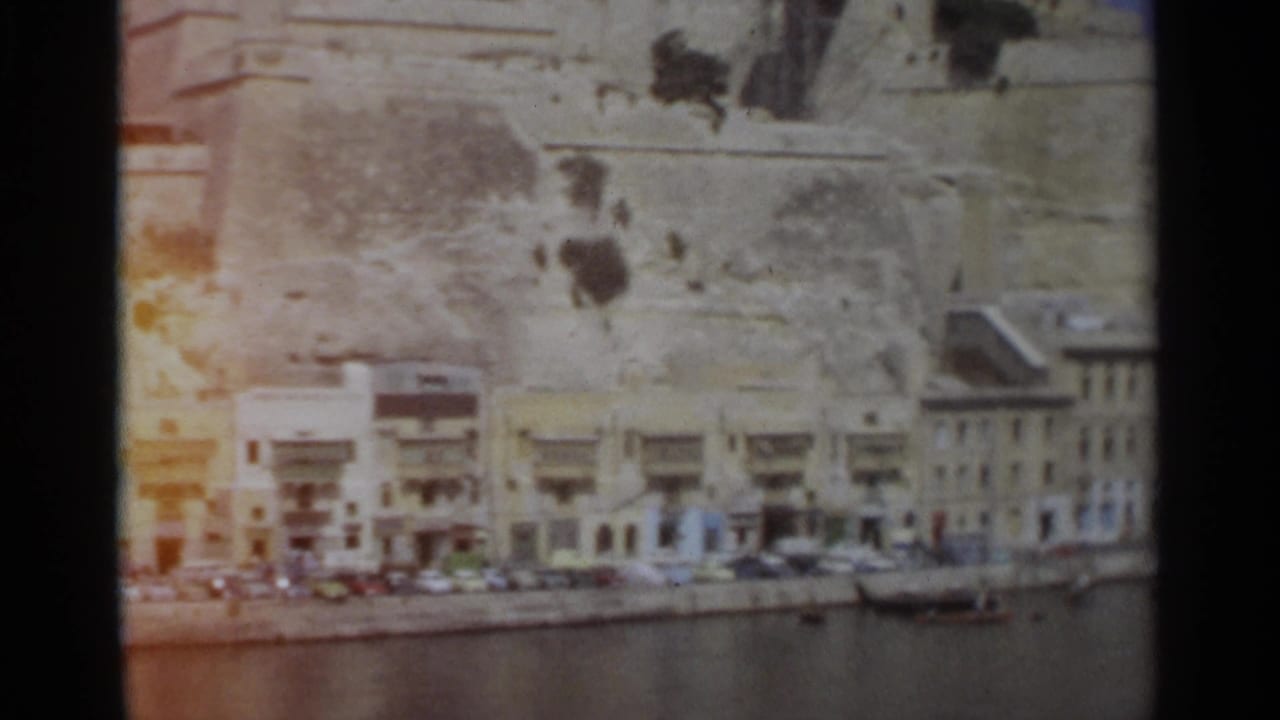 Faded 8mm frame of the Tangier waterfront, with parked cars, a small boat, pale buildings, and a reddish light leak at the film edge.