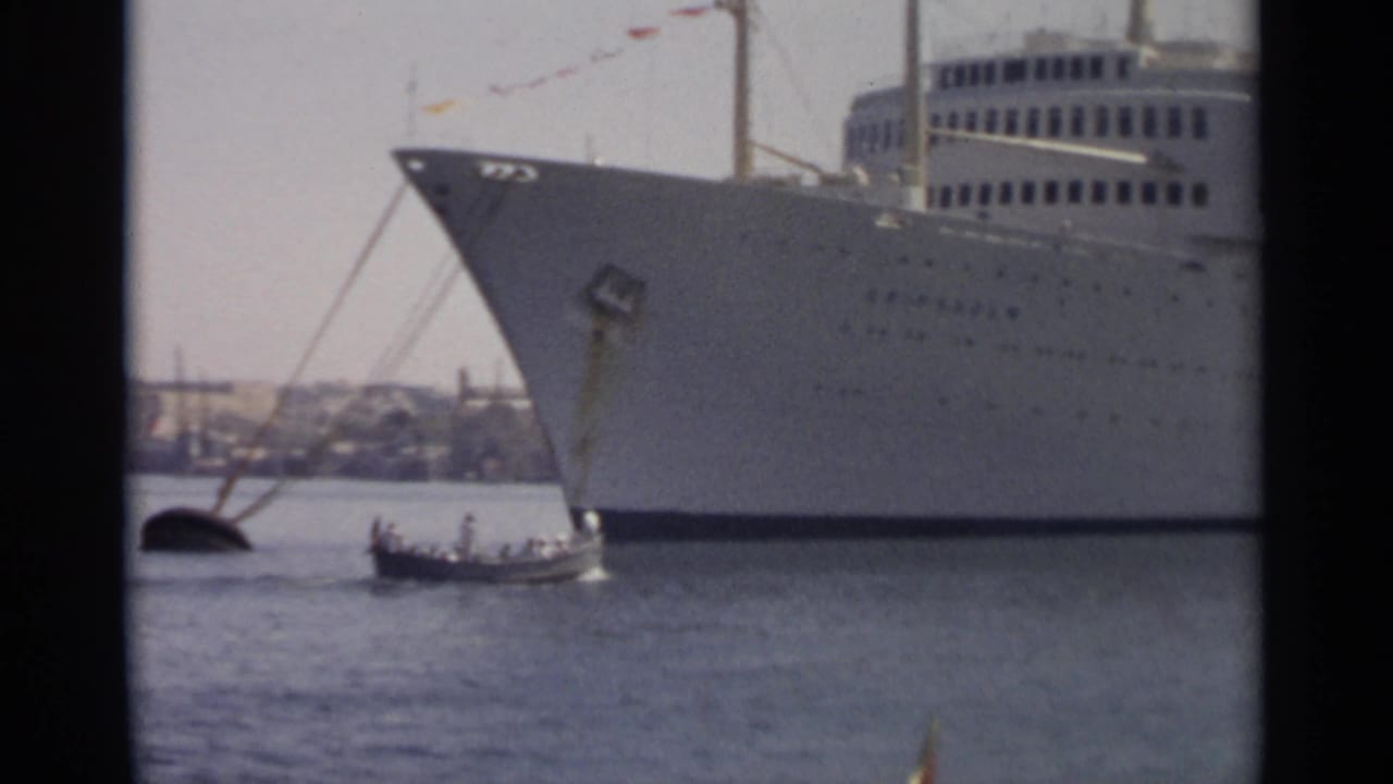 1965 Tangier harbor frame showing the bow of a large white ocean liner with a small boat passing nearby on the water.