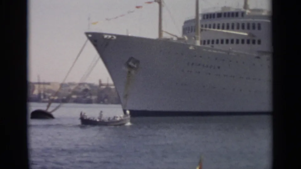 1965 Tangier harbor frame showing the bow of a large white ocean liner with a small boat passing nearby on the water.