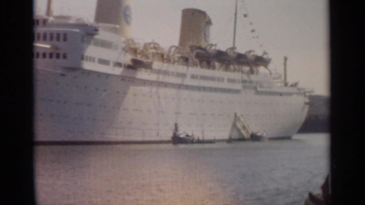 1965 Tangier harbor frame showing the side of a white ocean liner with lifeboats and small boats at the waterline.
