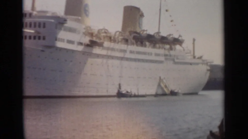 1965 Tangier harbor frame showing the side of a white ocean liner with lifeboats and small boats at the waterline.