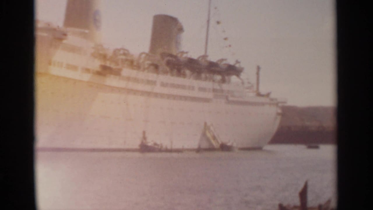 1965 Tangier harbor view with a large white ocean liner, small boats on the water, and shoreline in soft 8mm color.