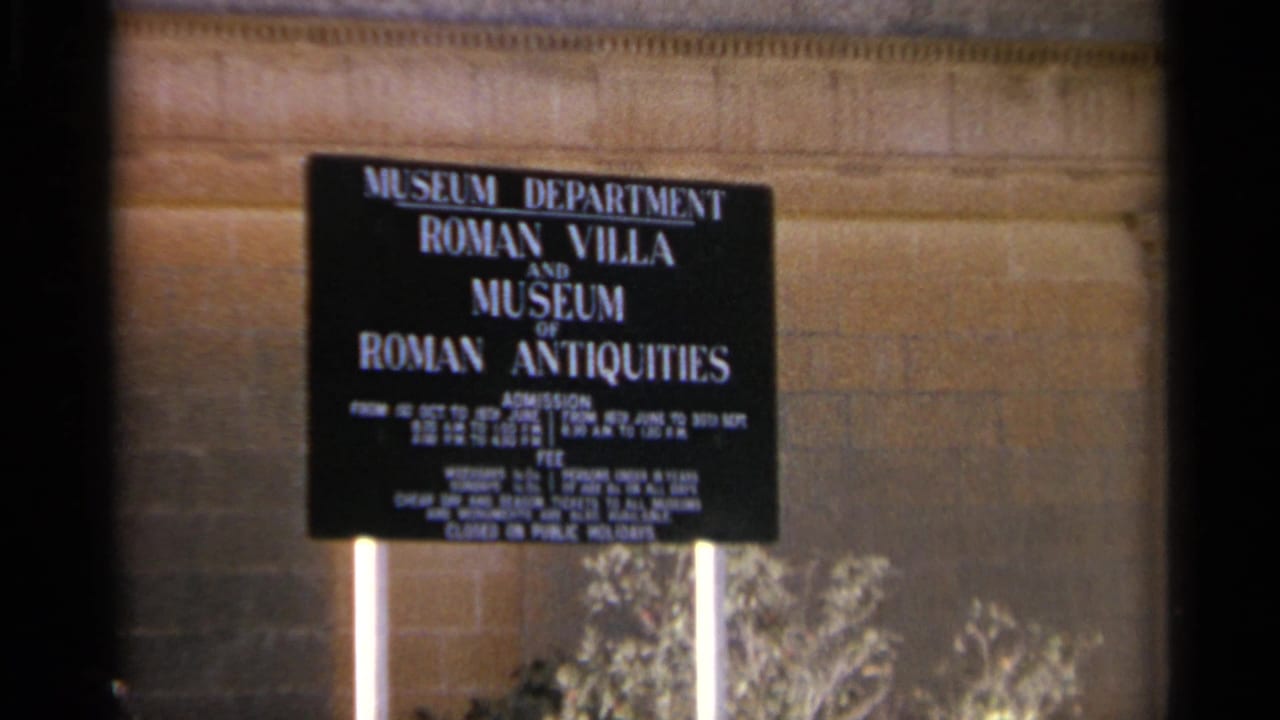 1965 Tangier museum sign reading Roman Villa and Museum of Roman Antiquities, mounted before a stone wall with plants below.