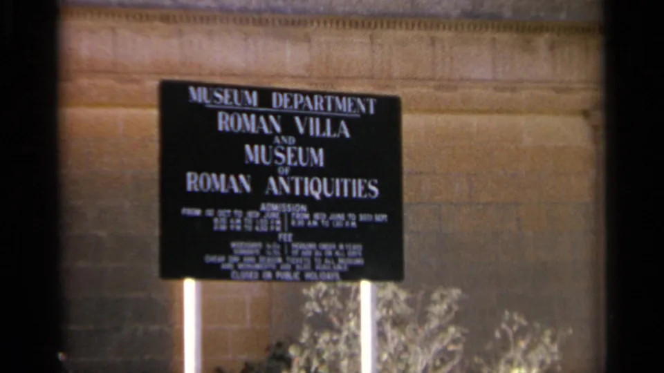 1965 Tangier museum sign reading Roman Villa and Museum of Roman Antiquities, mounted before a stone wall with plants below.