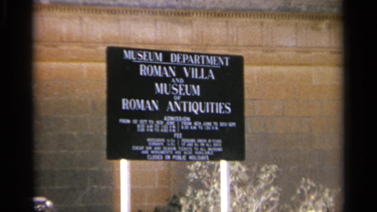 Tangier Roman antiquities museum sign held in frame, with white posts, a tan stone wall, and low vegetation visible below.