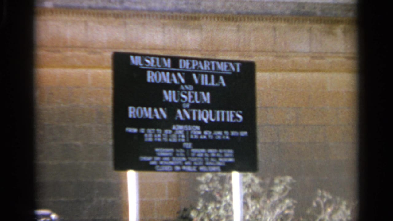 Black museum sign on a stone wall with white supports and shrubs below.