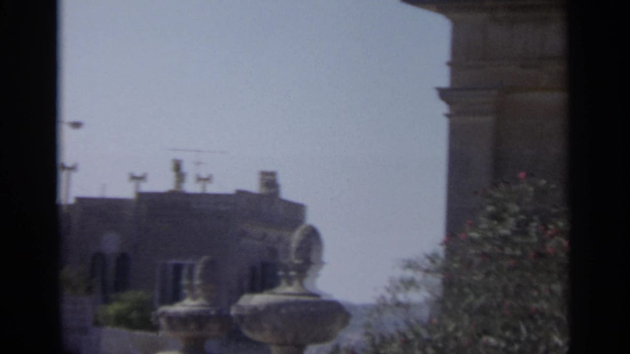 Handheld 8mm view of Tangier exterior architecture with stone railing forms, rooftop silhouettes, vegetation, and washed blue sky.