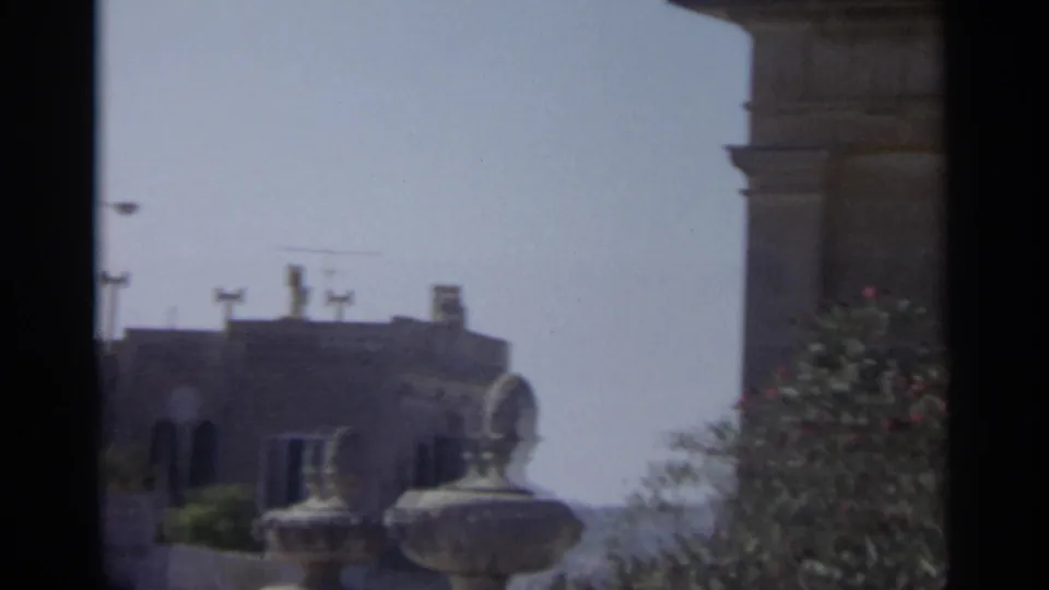 Handheld 8mm view of Tangier exterior architecture with stone railing forms, rooftop silhouettes, vegetation, and washed blue sky.