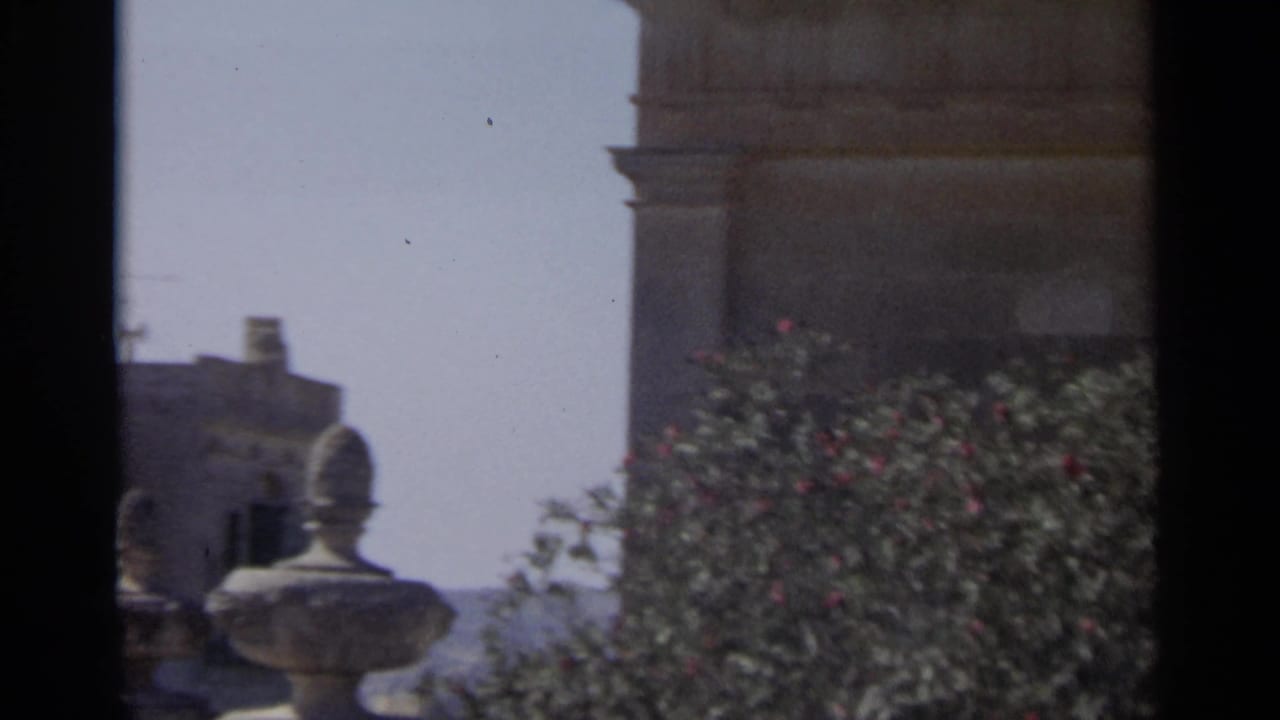 1965 Tangier home-movie frame showing a stone facade, leafy shrubs, and a decorative urn-like form near the edge of the building.