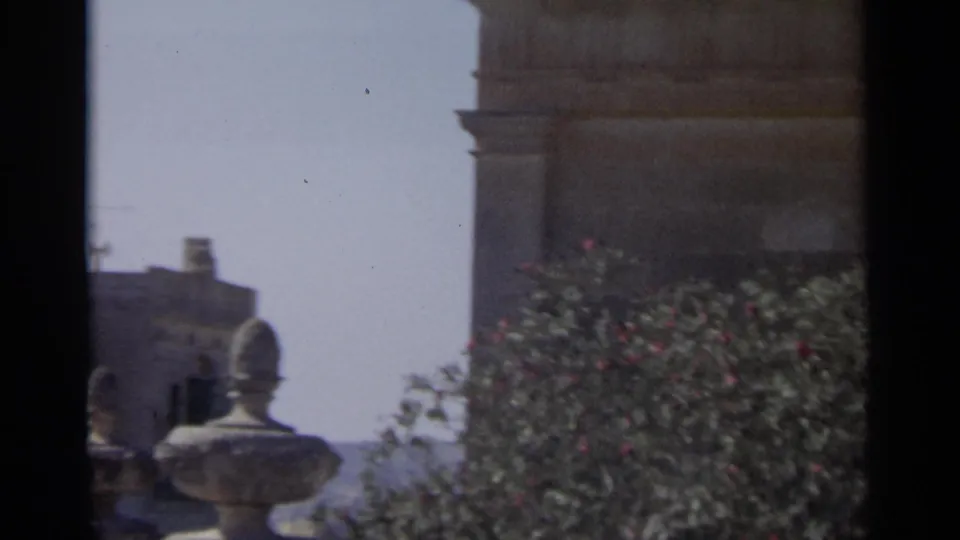 1965 Tangier home-movie frame showing a stone facade, leafy shrubs, and a decorative urn-like form near the edge of the building.