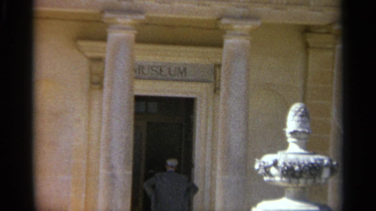 1965 Tangier home-movie frame shows a man in a light hat and dark jacket stepping from a museum doorway into bright sun.