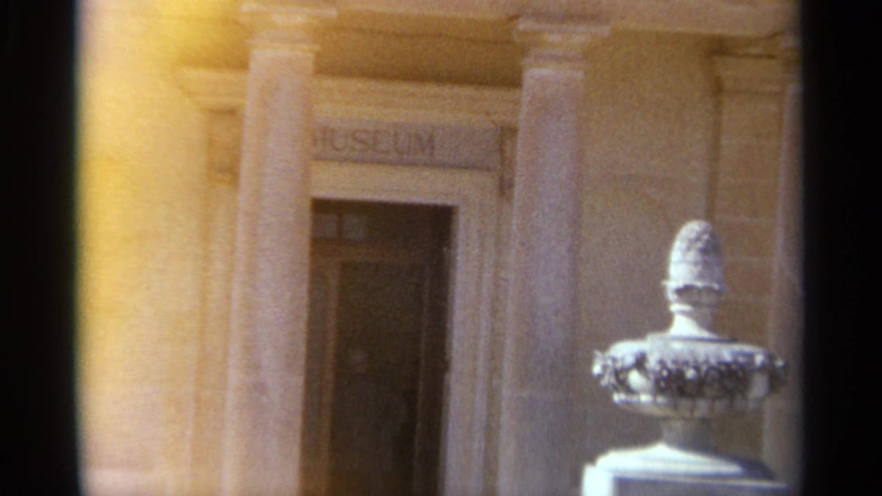 Warm 1965 Tangier 8mm frame of a museum entrance with stone columns, carved museum sign, open doorway, and white urn sculpture.