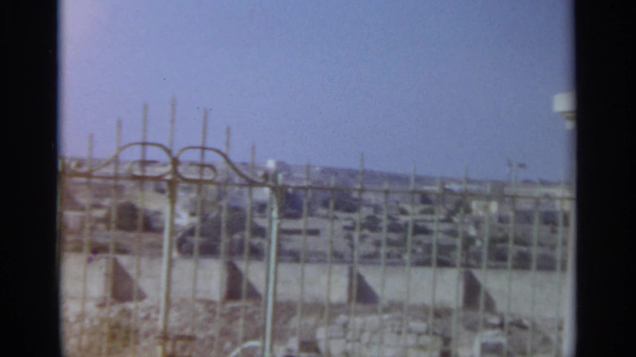 1965 Tangier 8mm frame looking through a metal gate toward a sunlit hillside cityscape beneath a clear blue sky.