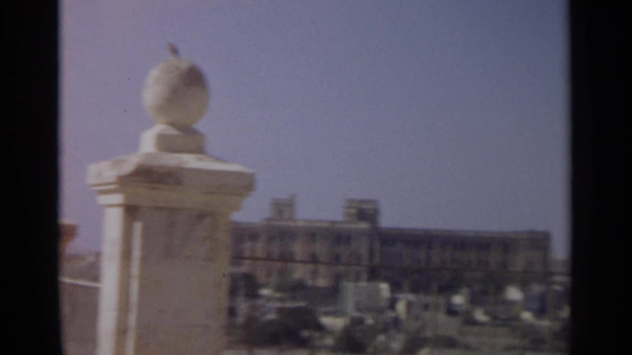 1965 Tangier home-movie frame with a pale stone finial in the foreground and soft distant buildings beyond a vertical dark edge.