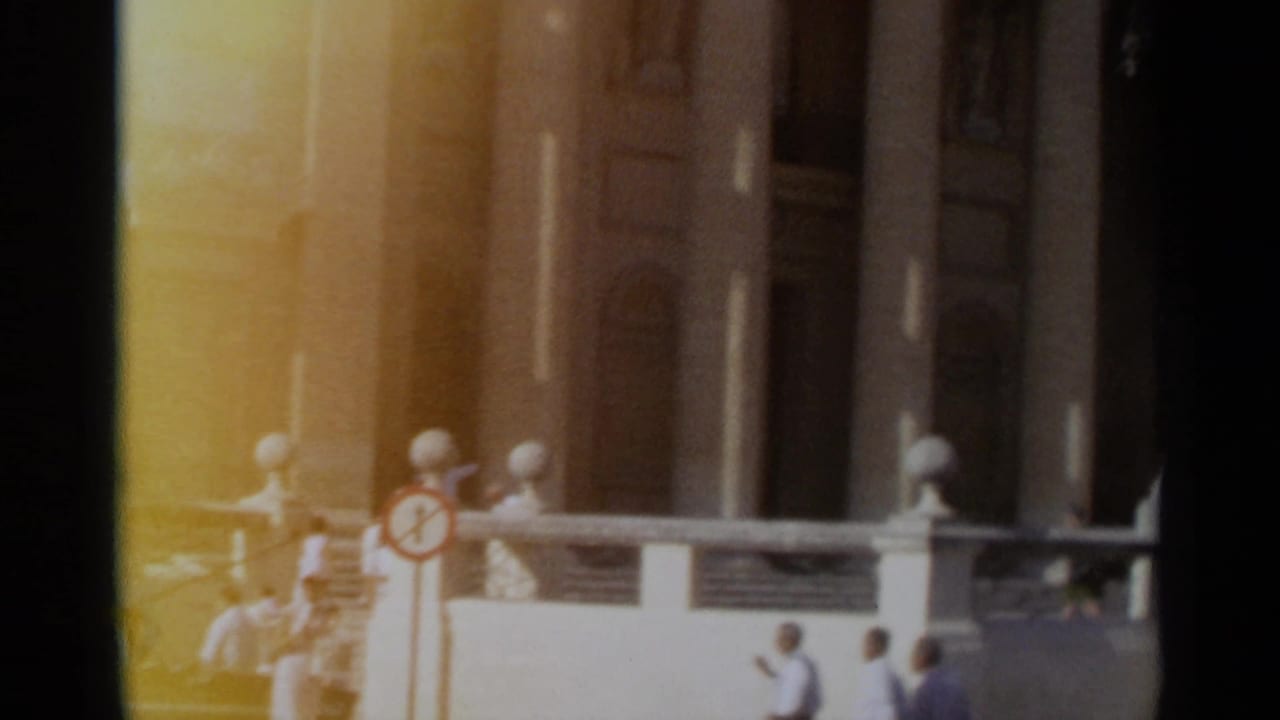 1965 Tangier 8mm frame showing a columned building frontage, balustrade, small pedestrians, and a parked car in strong sunlight.