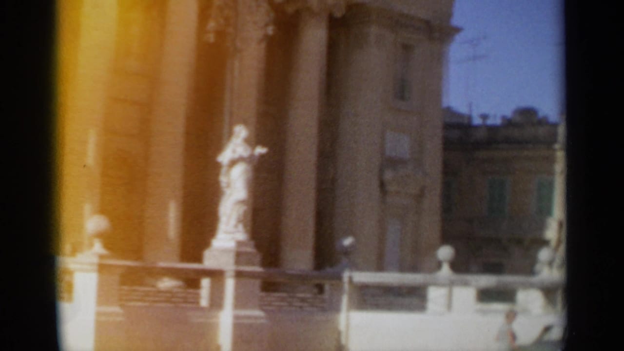 A Tangier 1965 home-movie frame shows a statue and columned exterior with a neighboring facade and blue sky beyond.