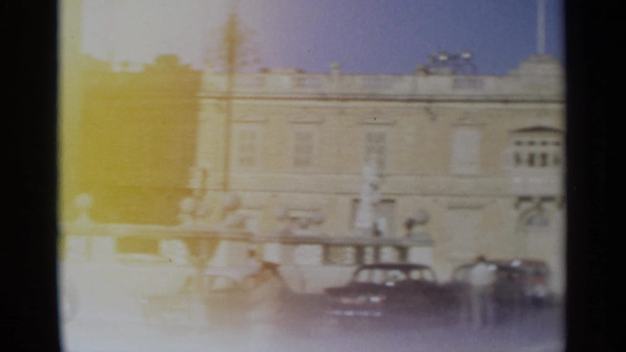 1965 Tangier home-movie frame showing parked cars and distant adults near a bright classical building facade.