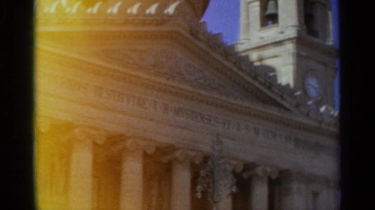 1965 Tangier home-movie frame showing a classical building facade with columns, roofline detail, and a partial clock tower under warm flare.